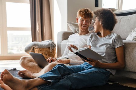 Photo for Young multinational couple using laptop and reading book while resting at home - Royalty Free Image
