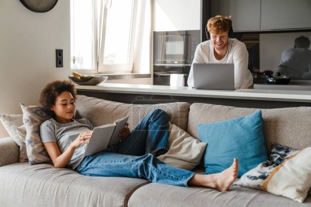 Foto de Joven pareja multinacional usando laptop y libro de lectura mientras descansa en casa - Imagen libre de derechos