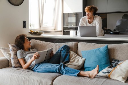Téléchargez les photos : Jeune couple multinational utilisant un ordinateur portable et un livre de lecture tout en se reposant à la maison - en image libre de droit