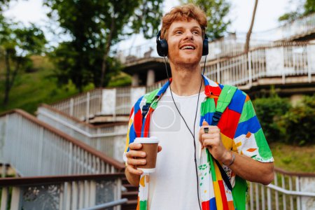 Photo for Portrait of young handsome smiling happy redhead man in headphones and colorful shirt holding coffee and looking aside, while standing on the street - Royalty Free Image