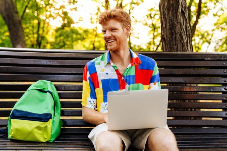 Photo for Young handsome smiling redhead man with laptop looking aside, while sitting with backpack on bench in park - Royalty Free Image