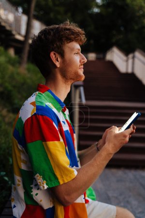 Photo for Side portrait of young handsome calm man holding phone and looking aside , while sitting on bench in park - Royalty Free Image