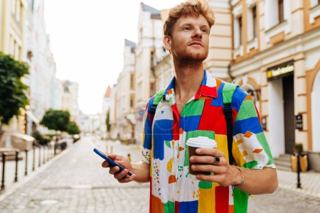 Photo for Portrait of young handsome calm redhead man with phone and coffee looking aside, while standing on the street - Royalty Free Image