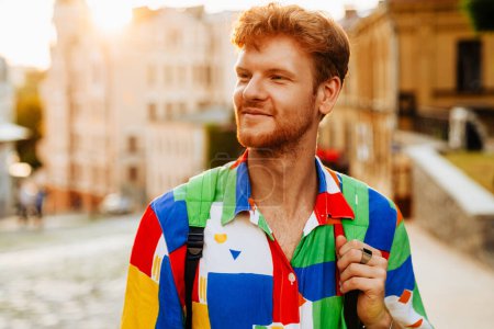 Photo for Portrait of young handsome calm redhead man in colorful shirt looking aside, while standing in the sun on the street - Royalty Free Image