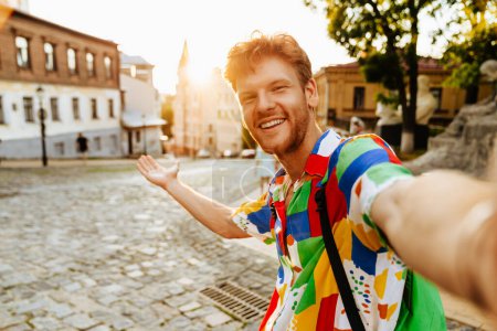 Foto de Selfie de joven guapo pelirrojo sonriente en camisa colorida con la palma levantada de pie en la calle soleada - Imagen libre de derechos