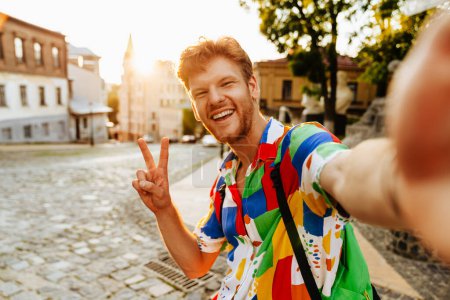 Photo for Selfie of young handsome smiling redhead man showing victory gesture with fingers, whuile standing on the street - Royalty Free Image