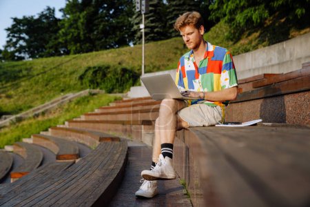 Téléchargez les photos : Jeune homme élégant et concentré travaillant avec un ordinateur portable, assis avec les jambes croisées sur tribune dans le parc - en image libre de droit