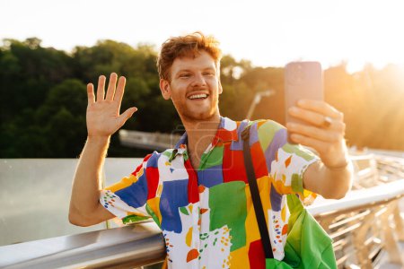 Photo for Young handsome stylish smiling man in colorful shirt with raised hand waving and taking selfie on his phone, while standing leaning on railing in the sun - Royalty Free Image