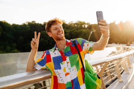 Photo for Young handsome stylish smiling man in colorful shirt showing victory gesture with fingers and taking selfie on his phone, while standing leaning on railing in the sun - Royalty Free Image