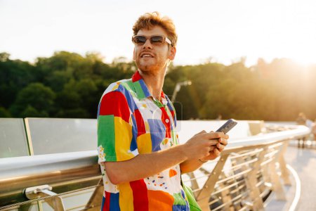 Photo for Young handsome stylish smiling man in sun glasses holding phone and looking aside , while standing leaning on railing in the sun - Royalty Free Image
