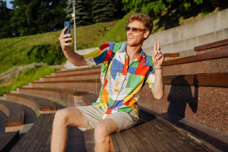 Téléchargez les photos : Jeune homme élégant et souriant dans des lunettes de soleil prenant selfie au téléphone et montrant un geste de victoire, assis sur tribune dans le parc - en image libre de droit