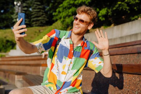 Téléchargez les photos : Jeune homme élégant et souriant dans des lunettes de soleil prenant selfie au téléphone et agitant la main levée, assis sur tribune dans le parc - en image libre de droit