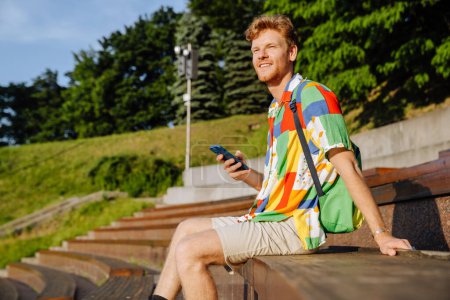 Téléchargez les photos : Jeune homme élégant et souriant avec téléphone regardant de côté, assis sur tribune à l'extérieur - en image libre de droit