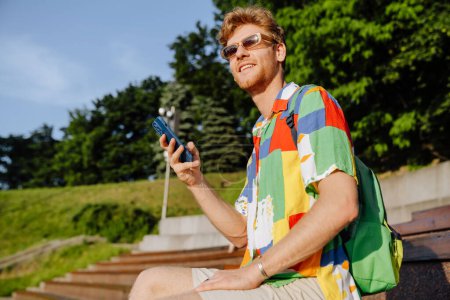Téléchargez les photos : Jeune homme élégant et souriant dans des lunettes de soleil avec téléphone regardant de côté, assis dans un parc ensoleillé - en image libre de droit