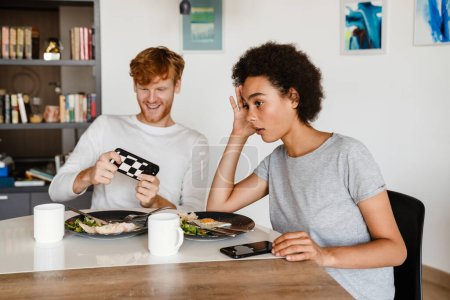 Photo for Young disappointed african woman propping her head and looking aside , while sitting near her smiling boyfriend who playing his phone during breakfast - Royalty Free Image