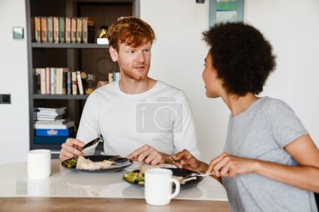 Téléchargez les photos : Jeune beau couple interracial se regardant et prenant le petit déjeuner ensemble à la maison - en image libre de droit