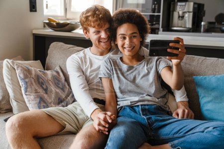Téléchargez les photos : Jeune couple interracial heureux prenant selfie avec téléphone ensemble, tout en tenant la main et assis sur le canapé dans le salon confortable à la maison - en image libre de droit