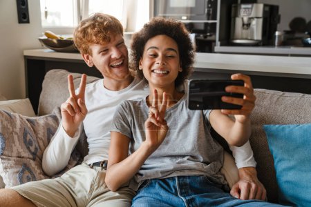 Téléchargez les photos : Jeune couple interracial heureux prendre selfie avec téléphone ensemble et faire geste de victoire avec les doigts, tout en étant assis sur le canapé dans le salon confortable à la maison - en image libre de droit