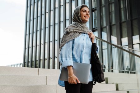 Foto de Joven mujer musulmana sonriente con pañuelo en la cabeza sosteniendo el ordenador portátil mientras está de pie en las escaleras con el edificio de vidrio en el fondo - Imagen libre de derechos
