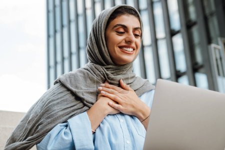 Téléchargez les photos : Femme musulmane portant foulard parlant par appel vidéo sur ordinateur portable et tenant la main à la poitrine tout en étant assis sur les escaliers à l'extérieur - en image libre de droit