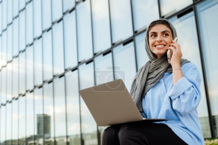 Fröhliche Muslimin mittleren Alters im Hijab, die auf dem Handy spricht und Laptop benutzt, während sie draußen auf der Straße der Stadt sitzt