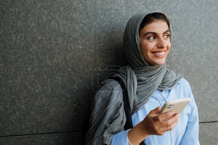 Foto de Sonriente mujer musulmana de mediana edad usando un teléfono inteligente y mirando a un lado mientras está al aire libre sobre la pared gris - Imagen libre de derechos