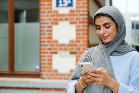 Téléchargez les photos : Femme musulmane d'âge moyen portant hijab à l'aide d'un smartphone tout en restant debout à l'extérieur dans la rue de la ville - en image libre de droit