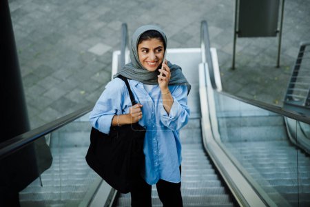 Foto de Mujer musulmana joven con pañuelo en la cabeza usando el teléfono celular mientras está de pie en escaleras mecánicas al aire libre - Imagen libre de derechos