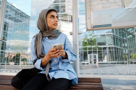 Téléchargez les photos : Jeune femme musulmane portant un foulard à l'aide d'un téléphone portable assis sur l'arrêt de bus à l'extérieur - en image libre de droit