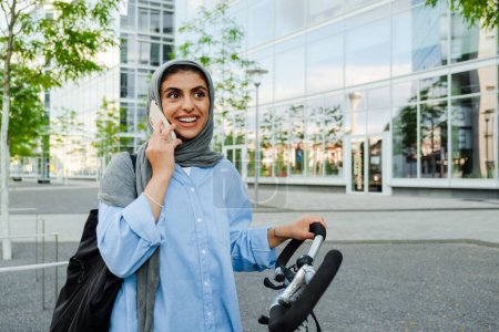 Foto de Joven mujer de negocios musulmana con pañuelo en la cabeza usando el teléfono celular mientras está de pie en bicicleta al aire libre - Imagen libre de derechos