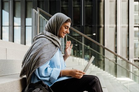 Photo for Young muslim woman wearing headscarf using laptop while sitting on stairs outdoors - Royalty Free Image