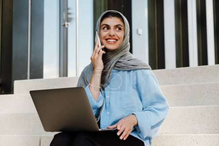 Téléchargez les photos : Jeune femme musulmane portant le foulard à l'aide d'un téléphone portable et ordinateur portable tout en étant assis sur les escaliers à l'extérieur - en image libre de droit