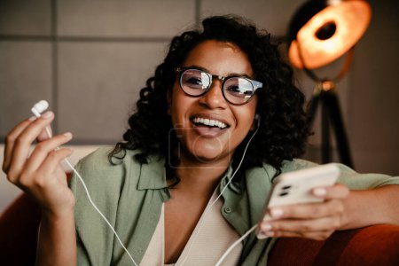 Photo for Cheerful young african american woman in eyeglasses putting on earphones while using smartphone at home - Royalty Free Image