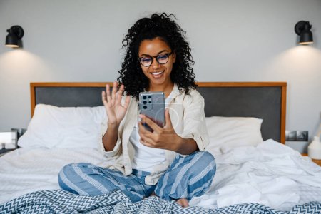 Photo for Black young woman in eyeglasses using mobile phone while resting on bed at home - Royalty Free Image