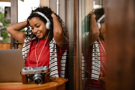 Téléchargez les photos : Jeune femme noire dans les écouteurs en utilisant un ordinateur portable tout en se reposant dans le café à l'intérieur - en image libre de droit