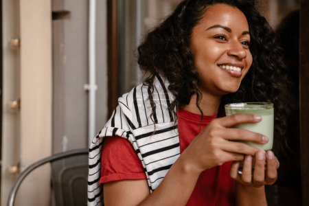 Téléchargez les photos : Jeune femme noire souriant et buvant matcha latte tout en étant assis au café à l'extérieur - en image libre de droit