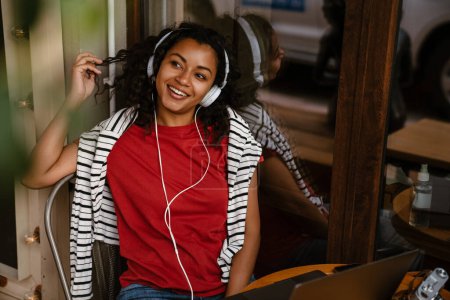 Téléchargez les photos : Jeune femme noire écouter de la musique tout en travaillant avec ordinateur portable au café à l'extérieur - en image libre de droit