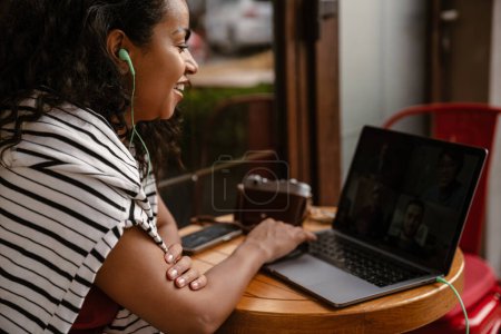 Téléchargez les photos : Jeune femme noire dans les écouteurs de travail avec ordinateur portable au café à l'extérieur - en image libre de droit
