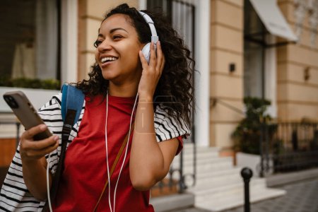 Téléchargez les photos : Jeune femme noire dans les écouteurs en utilisant un téléphone mobile tout en se tenant à l'extérieur - en image libre de droit