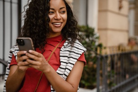 Téléchargez les photos : Jeune femme noire utilisant un téléphone portable tout en restant debout à l'extérieur du bâtiment - en image libre de droit