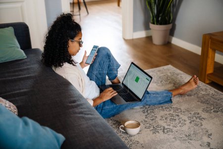 Black young woman using gadgets and drinking coffee while sitting on floor at home