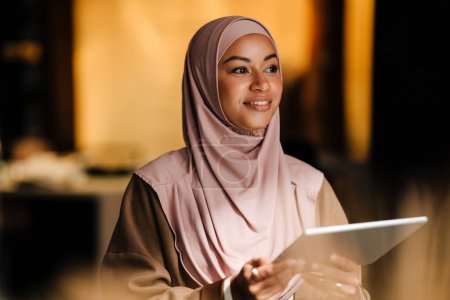 Téléchargez les photos : Arabe femme portant hijab travailler sur tablette tout en se tenant à l'intérieur dans le bureau moderne - en image libre de droit