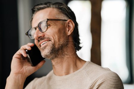Téléchargez les photos : Gros plan de l'homme blanc d'âge moyen dans les lunettes souriant tout en parlant sur téléphone portable à l'intérieur - en image libre de droit