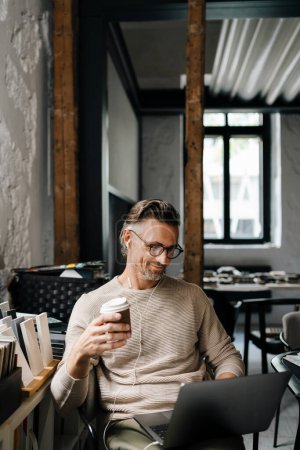 Photo for White middle-aged man in earphones drinking coffee while using laptop computer in modern office - Royalty Free Image