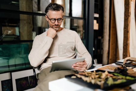 Téléchargez les photos : Pensive homme d'âge moyen blanc dans les lunettes de travail avec tablette informatique à l'atelier - en image libre de droit