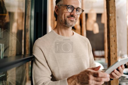 Téléchargez les photos : Blanc gai homme d'âge moyen dans les lunettes à l'aide d'un ordinateur tablette tout en travaillant dans le bureau moderne - en image libre de droit