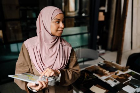 Téléchargez les photos : Femme arabe souriante portant hijab travaillant sur tablette tout en se tenant debout dans le bureau moderne - en image libre de droit