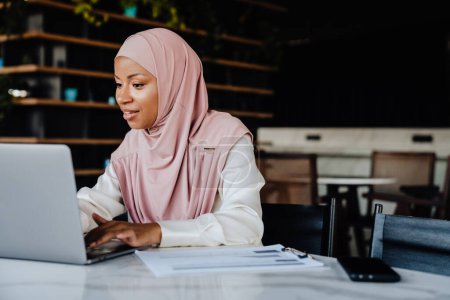 Foto de Mujer musulmana joven con pañuelo en la cabeza sonriendo mientras trabaja en el ordenador portátil en la oficina - Imagen libre de derechos