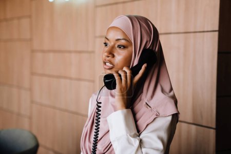 Foto de Joven empresaria musulmana hablando por teléfono mientras trabaja en recepción - Imagen libre de derechos