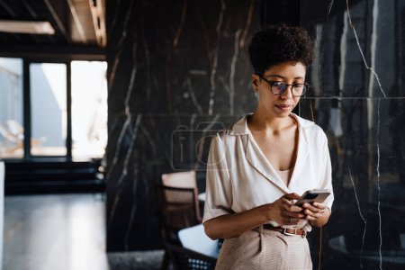 Téléchargez les photos : Heureuse femme d'affaires noire utilisant un téléphone portable tout en travaillant dans le hall du bureau - en image libre de droit